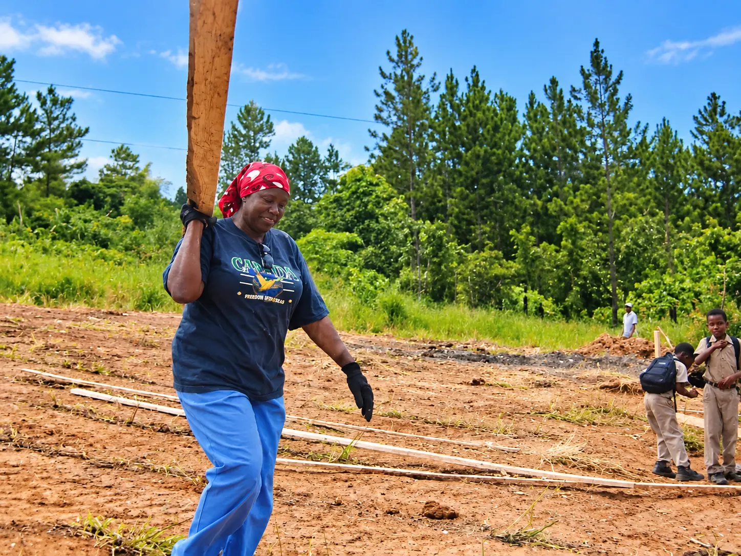 A woman in a dark blue t-shirt carries a long timber on her shoulder across a cleared red-dirt building site, with two schoolchildren in beige uniforms standing nearby and a wooded hillside behind