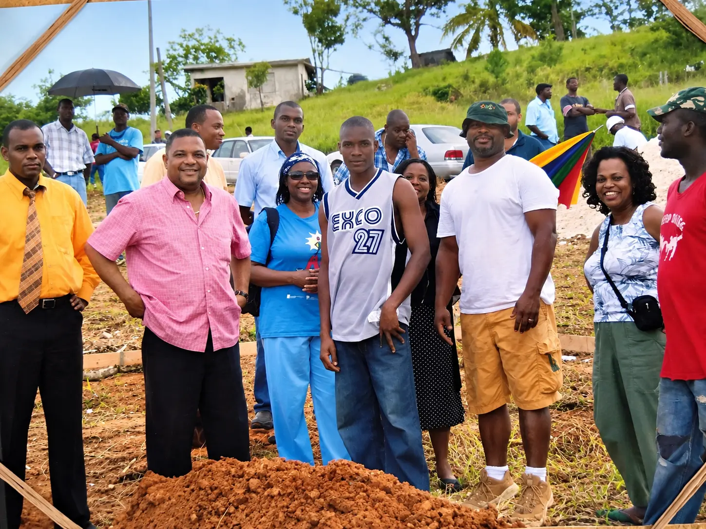 A community group of about nineteen people pose behind a fresh mound of red clay at a rural building site, with parked cars, a small concrete house and a grassy hillside behind