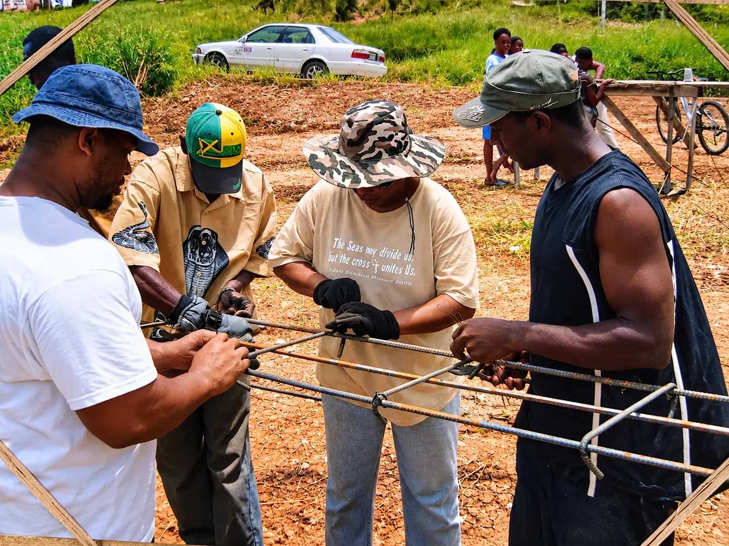 Four workers assemble and wire-tie a rectangular rebar cage on red dirt at a construction site, with a white sedan, bicycles and a wooden work area in the background