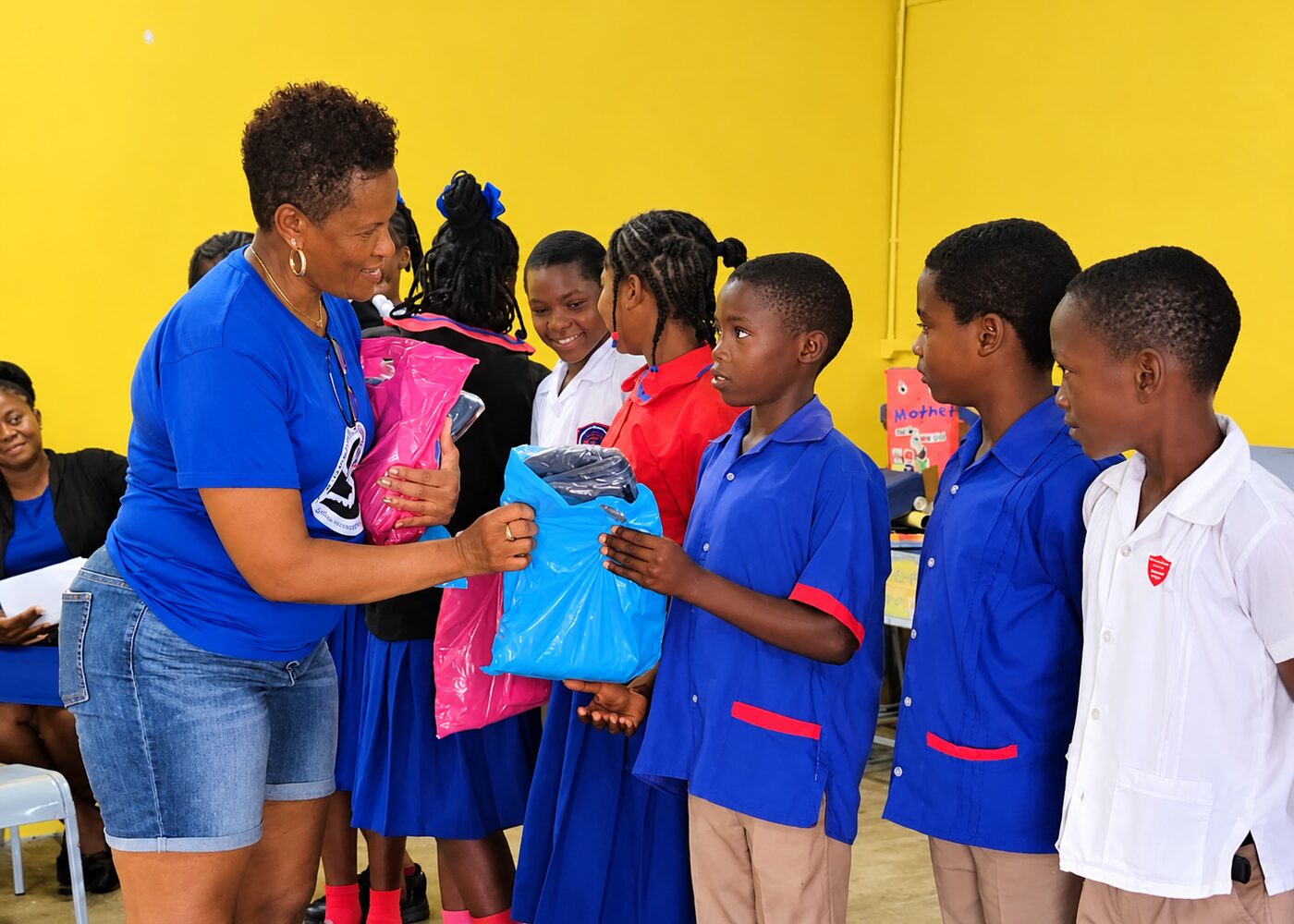 A woman in a blue t-shirt hands a bright blue gift bag to a smiling schoolboy while a line of children in school uniforms waits behind him in a room with yellow walls