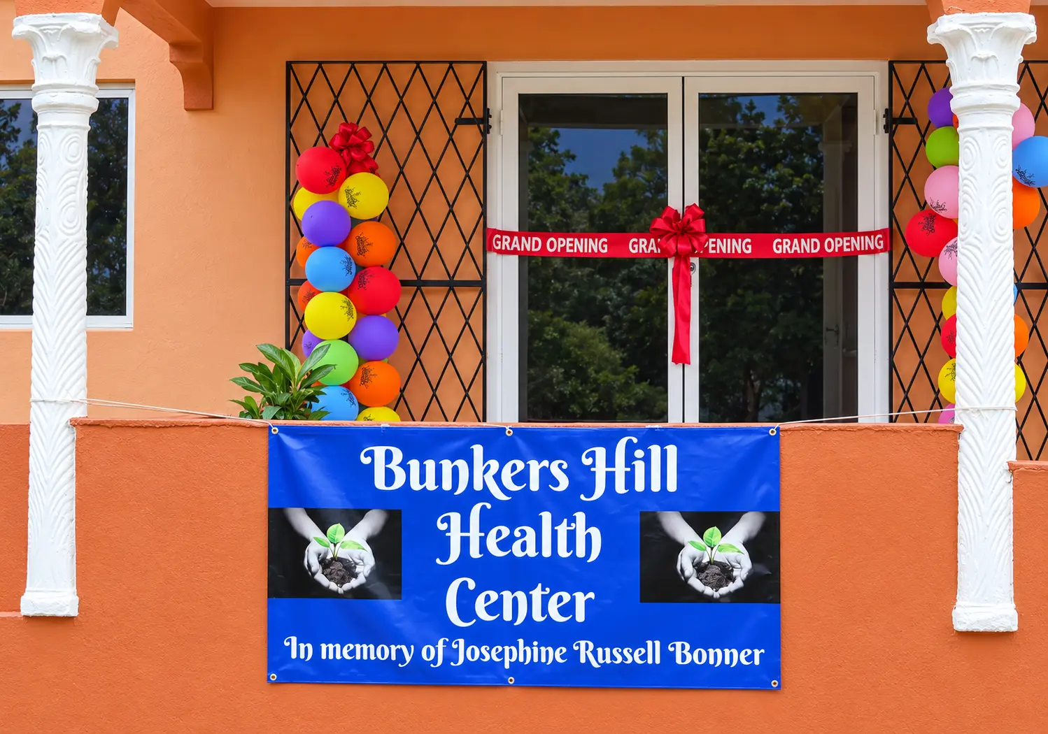 The entrance of the Bunkers Hill Health Center decorated for its opening — a red GRAND OPENING ribbon with a bow across the glass double doors, towers of colourful balloons either side, and a royal blue banner on the porch wall reading Bunkers Hill Health Center, in memory of Josephine Russell Bonner