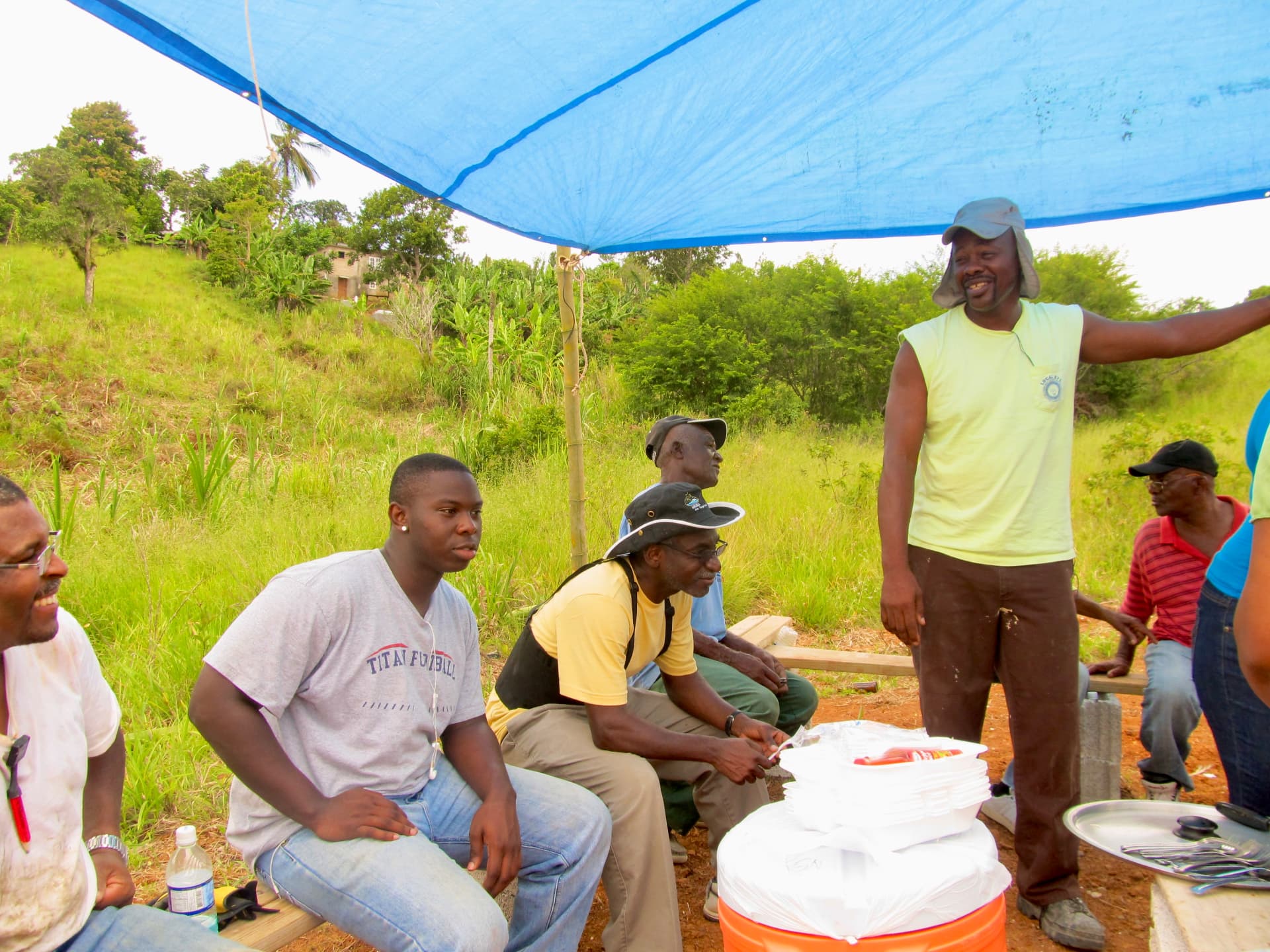 A group of volunteers and community members gathered beneath a blue tarp canopy on a grassy hillside, some seated on a wooden bench and one standing in a sleeveless shirt and sun cap, with an orange cooler and stacked white food containers in the foreground