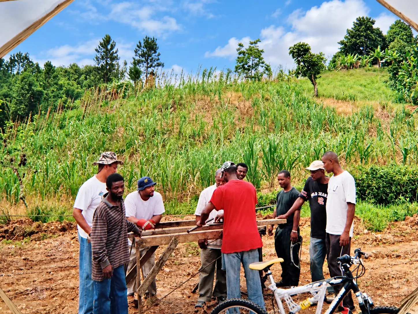 Nine men gather around a wooden workbench on a cultivated hillside, one bending to work rebar with a pipe tool while a mountain bike rests in the foreground