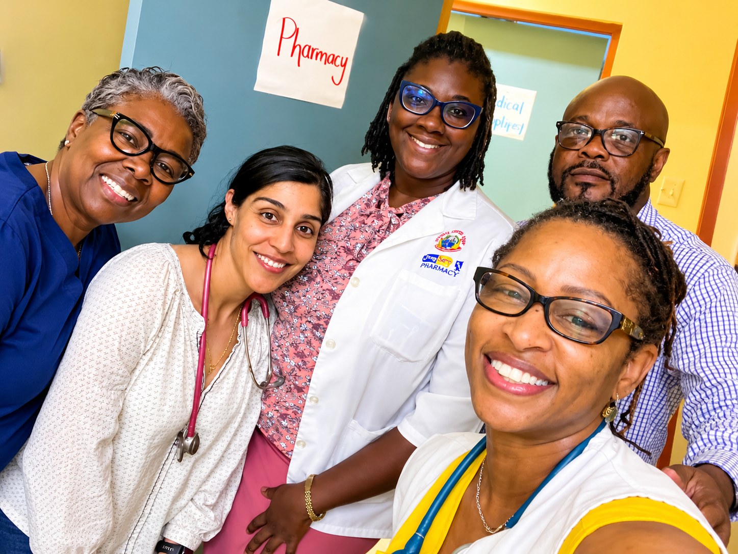 Five health professionals smiling together inside a clinic, with a handwritten Pharmacy sign on the wall behind them