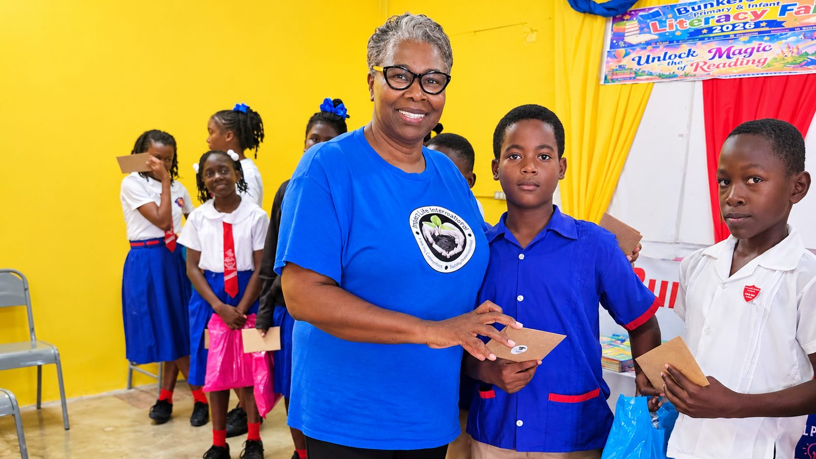 A smiling woman in a blue t-shirt hands a paper envelope to a schoolboy in a blue uniform while children in school uniforms wait in line in a room with yellow walls and a Literacy Fair 2026 banner