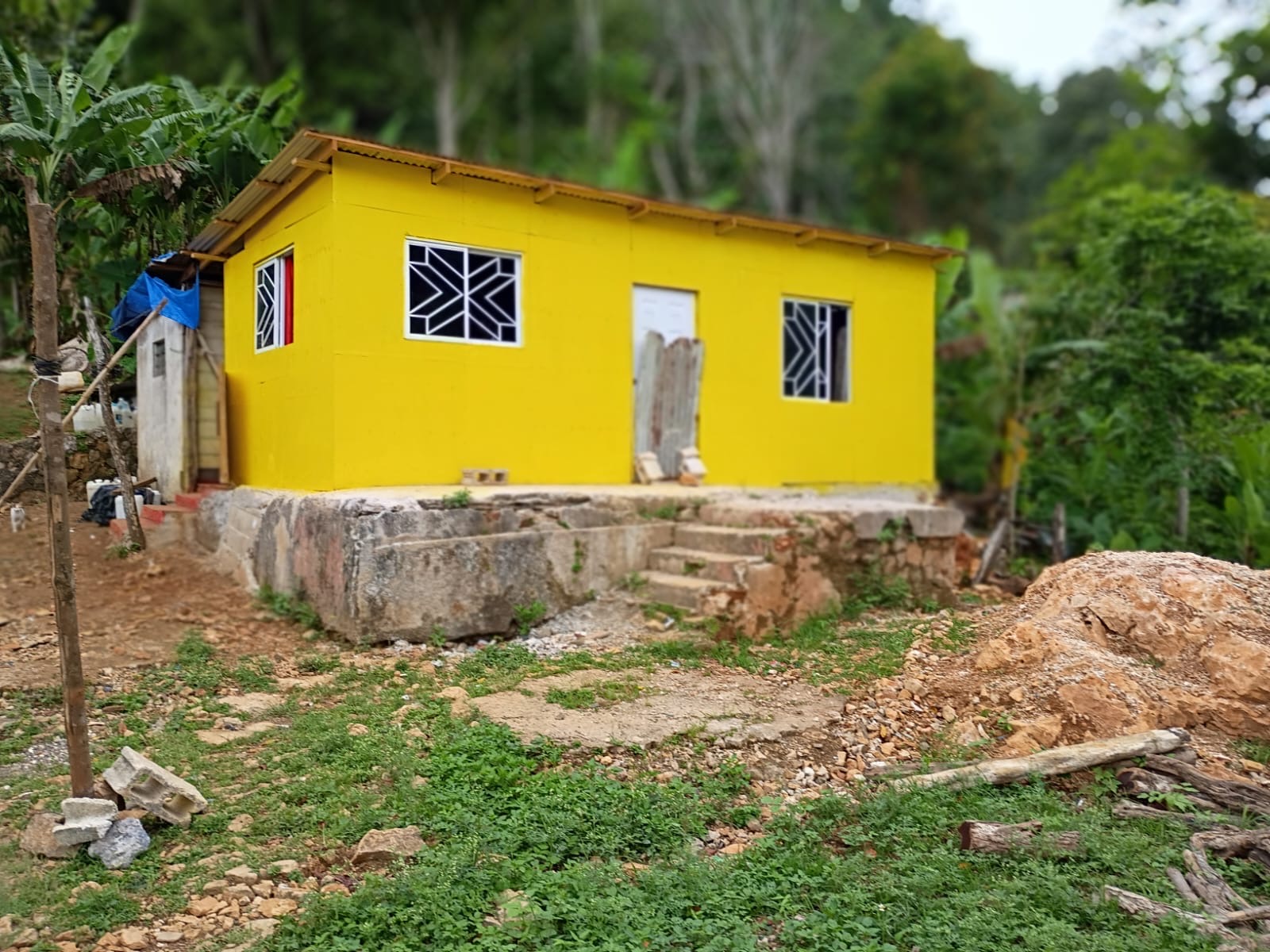 A yellow single-story house in Jamaica standing on a rough concrete-block foundation, its corrugated metal roof and white-barred windows in place, with a blue tarp covering an adjoining structure and loose earth, logs and masonry blocks in the yard at the foot of a wooded hillside
