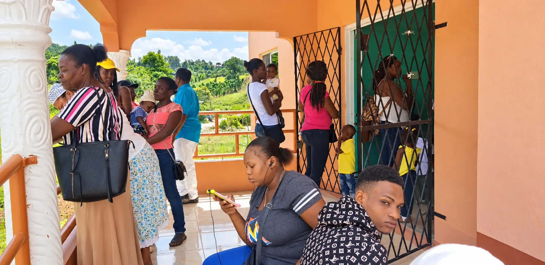 A group of adults, children and babies gathered on the covered porch of a peach-coloured building with an orange railing and a green door behind a metal security gate, while a woman seated in front looks at her phone and a man in a turquoise shirt leans on the railing overlooking green hills